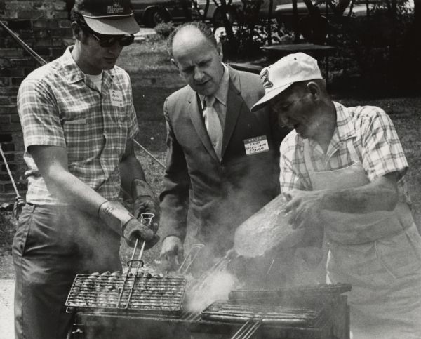 A man in a suit is watching as one man is pouring liquid onto a grill, while another man on the left is holding a grill holder full of brats. Caption reads: "<b>EXPERT EYE</b> - Fire Chief William Stamm kept an eye on things Thursday as Ald. John Czarnezki (left) and Ralph Wroblewski (right) grilled bratwurst at the annual aldermen's icnic [sic] at Veteran's Park, 5607 S. 6th St. Wroblewski is an employe of the city Forestry Department."