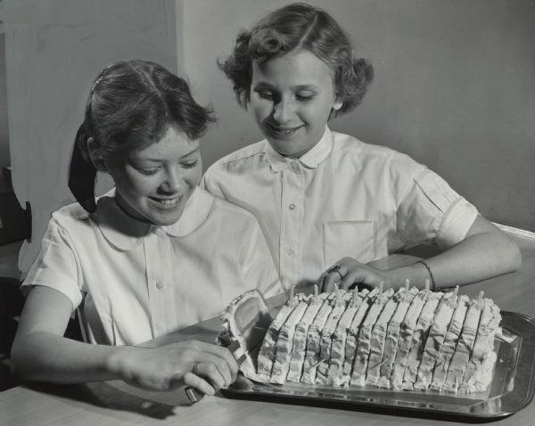 Two girls are smiling and looking at a loaf of sliced and apparently frosted food. Caption reads: "<b>A bread loaf filled</b> with peanut butter and topped with cream cheese was inspected by Mardene Dreger (left), 11, of 505 N. 106th St., and Janice Hasselstrom, 12, of 336 N. 113th St., at a food festival at Underwood school, 11132 W. Potter Rd., Wauwatosa. The theme of the festival was taken from a Wisconsin state department of health booklet. The party stressed orange juice, cakes of bread and cheese, and other nutritious dishes instead of the customary sweets."