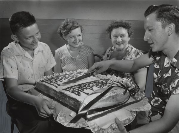Three young people and one adult woman are sitting around a cake shaped like a book with writing on its open pages. One young man is holding a knife over the cake. Caption reads: "<b>Graduates of the Hays school,</b> 2431 S. 10th st., had their names written in frosting on a cake served at a party for them at the school Wednesday. From left are James Lemke, 2566 S. 9th Pl.; Louise Mrozinski, 1017 W. Lincoln Av.; Miss Hester Mehl, teacher of the class, and Joseph Bartkiewicz, 1409 W. Harrison Av., the class president."