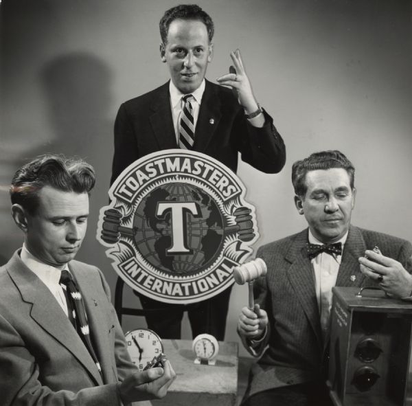A man in the center is standing behind a sign for Toastmasters International with one hand raised up. Two other men are sitting and looking at pocket watches, and the man on the right is also holding a gavel. Caption reads: "When a member of the Toastmasters club is given five minutes in which to make a speech he knows that five minutes is all he will get. If he tries to go over that the gavel will fall and he'll have to shut up, even if he is in the middle of a sentence. To illustrate this we have Wayne Linderman, 2227 S. 106th St., acting as timekeeper; Harry A. Schopler, 150 E. Fairmount Av., as speaker, and Frank I. Spangler, 5271 N. Bay Ridge Av., as gavel wielder. The Wisconsin convention of the Toastmasters club will be held in Milwaukee Apr. 16. After the dinner at 3651 N. 27th St., six speakers will compete for the state title. The winner will go on to a regional contest and the winners of the regionals will compete at the annual convention in Los Angeles this fall. The speakers competing for the Wisconsin title will not know what their subjects are to be until 2 p.m. of the day of competition.