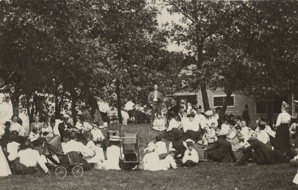 A man is standing on a small ridge, surrounded by people sitting on the ground. He is holding an open book and speaking. Caption reads: "Reuben G. Thwaites speaking, Homecoming of Newport, Wis., August, 1909."