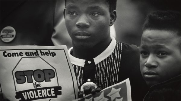 Two young men stand together. One is holding a sign that is barely visible. Next to him is another sign that reads "Come and help STOP the VIOLENCE". Caption reads: Antonio Holder (left), 13, and James Taylor, 14, listen during a youth rally on violence Friday at Martin Luther King Park. The rally and a march were part of "Stop the Violence" week, which encouraged alternatives to drug use, weapons and child abuse.