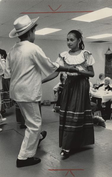 A young man and woman are dancing together on a stage. Other dancers and people are sitting at tables in the background. Caption reads: "The La Casa Senior Center at 410 Arcadian Ave., Waukesha, held a celebration Friday in honor of Mexican Independence Day. The event included dancing, music and a program that outlined the significance of the day. Silvia Quineonez, 9, and Dimas Ocampo, 9, both from St. Joseph's Church in Waukesha, performed a Mexican dance for the audience."