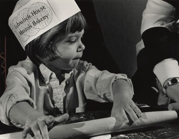 A young child is rolling dough with a rolling pin. She is wearing a paper hat that reads "Lubavitch House Matzoh Bakery." An adult. partially out of frame, is next to her on the right. Caption reads: "With Passover beginning on April 19, the Lubavitch House, 3109 N. Lake Dr., opened its bakery for groups to come and make their own traditional shmurah (watched) matzohs. According to a Lubavitch House spokeswoman, water must not contact the grain and flour used in shmurah matzohs from the time of harvest through the milling, sifting and packaging, or else the matzohs would not be kosher. Aviva Abergel, 3, of Milwaukee, learned how to roll out the unleavened dough to make matzoh."