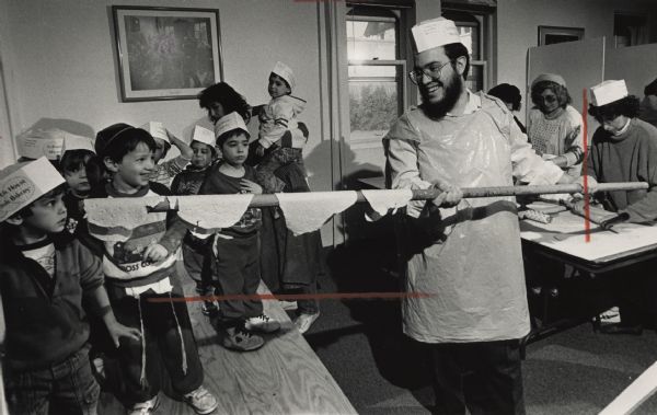 A man in an apron and baker's hat is smiling and holding a pole, with several rolled matzoh on it. On the left are several children wearing paper hats. At a table in the background on the right are several women rolling dough. Caption reads: "Yankie Fogel, a student from Brooklyn, N.Y., carried matzohs to the oven under the watchful eyes of several bakers."