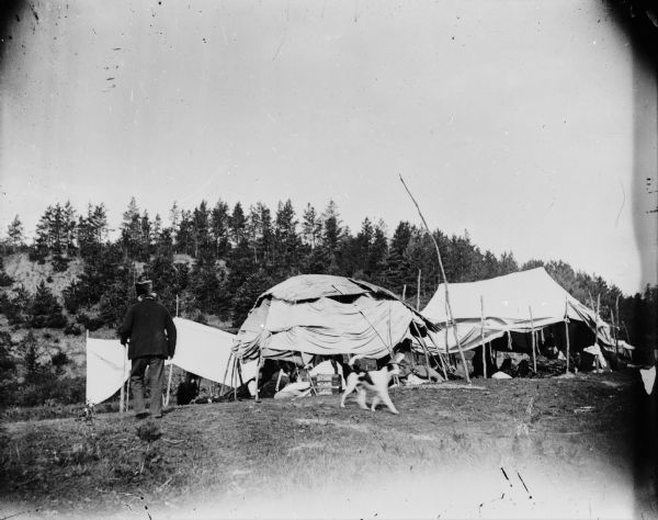 View of the Winnebago camp Old Monegar at Morrison Creek Bottoms. A man wearing a hat, and a black and white dog, are walking in front of the living shelters, a typical dwelling (chipoteke). There are people sitting inside. In the background is a tree-lined ridge.
