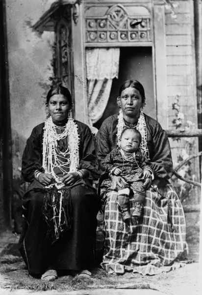 Studio portrait in front of a painted backdrop of two Indian (probably Winnebago) women. The woman on the right is holding a baby on her lap. The women are wearing white bead necklaces, bracelets, and silver drop earrings.