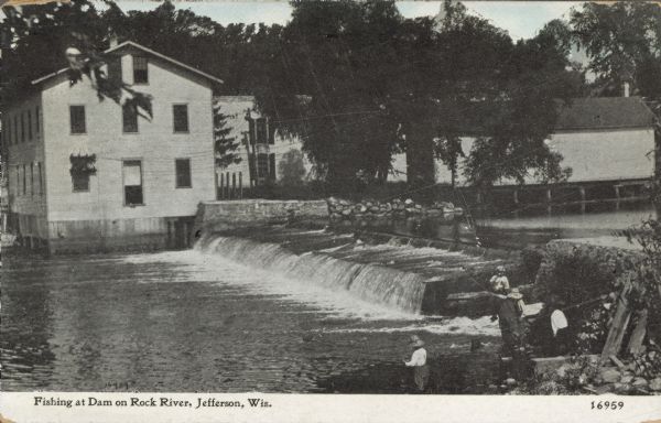 A group of boys are fishing on the river bank beside a dam. Buildings are on the far bank. Caption reads: "Fishing at Dam on Rock River, Jefferson, Wis."
