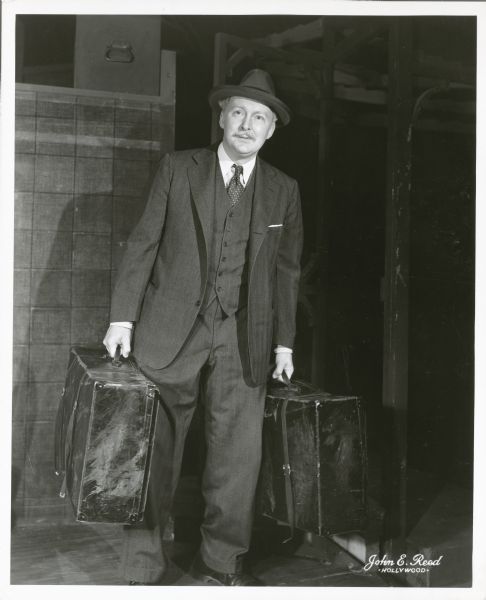 Albert Dekker, as Willy Loman, is standing and holding two large sample cases in the play "Death of a Salesman." He is dressed in a three-piece suit and is wearing a hat.