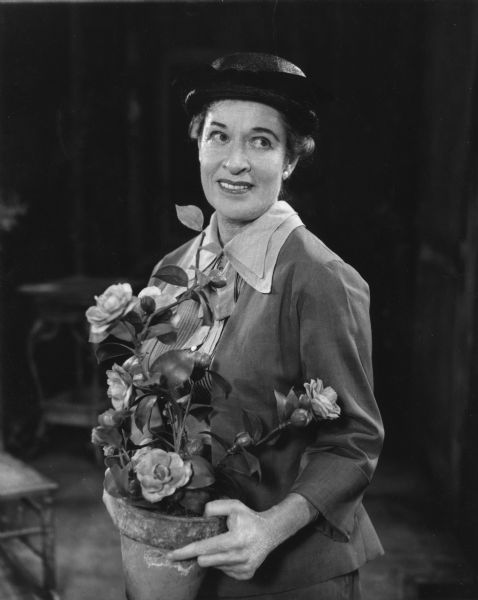 Anne Revere, as Anna Berniers in the play "Toys in the Attic," is holding a flower pot with large fake flowers. She is wearing a hat and is looking off to the side.
