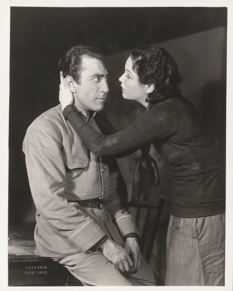 Herbert Biberman, as Terekhine, is sitting on a table and looking at an unidentified actress in a scene from the play "Red Rust." The actress has her hands on either side of his head and she is leaning towards him. He is dressed in a Bolshevik uniform.