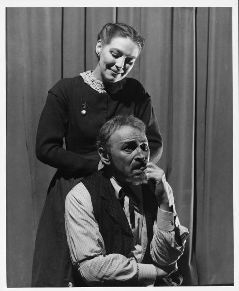 Sarah Cunningham is standing behind Morris Carnovsky, who is sitting, in a scene from the play "The World of Sholom Aleichem." She is smiling down on him while he is biting a finger and is looking worried.