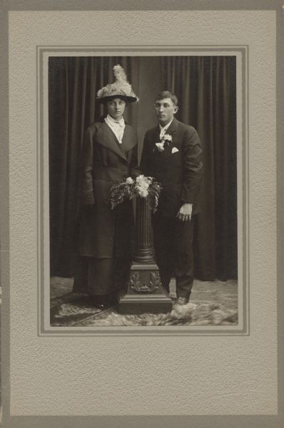 Full-length studio portrait of a woman and a man standing and posing together around a pedestal. The man is wearing a suit with a corsage of flowers, and the woman is wearing a dress, overcoat and feathered hat. She also appears to be holding a bouquet of flowers and ferns on top of the pedestal. Caption reads: "Frank Kreuser & Wife [Anna M. Kohl Kreuser]."