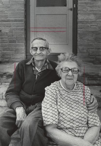 Portrait of a man and woman posing sitting on the steps in front of a door. The man has his arm around the woman's shoulder. Caption reads: "Archie and Dorothea Johnson live in the shadow of the Baraboo Bluffs."