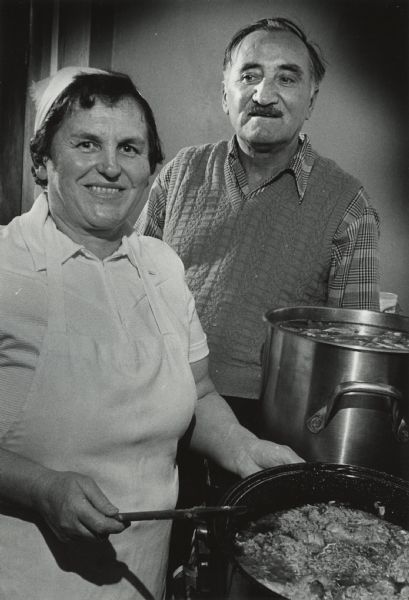 A woman and a man are standing next to two large pots of food on a stove. The woman is holding a utensil over one pot. Caption reads: "Dushanka and Gruja Bratkovich in their restaurant kitchen."