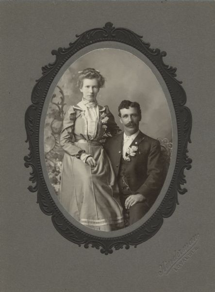 Oval-framed studio portrait in front of a painted backdrop of a woman and man posing sitting together. The woman is sitting on the arm of the chair, and is wearing a dress with a corsage of flowers. The man is wearing a suit with white bow tie, a vest and watch fob, and a flower boutonniere on the lapel of his jacket. Caption reads: "John & Belle Mingle (Mengel) family. To John Mengel from Sister Mamie."
