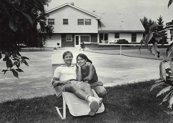 Portrait of a man and woman posing together on the lawn in front of a tennis court and a house. The man is sitting in a reclining lawn chair, and the woman is kneeling next to him. Caption reads: "Bob and Faye Becker have been together for more than half their lives."