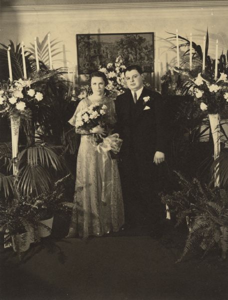 Portrait of a woman and a man standing and posing together. They are surrounded by ferns, flowers, and candles. The woman is wearing a dress, presumably a bridal gown, and is holding a bouquet of roses. The man is wearing a suit and tie, and a boutonniere. Caption reads: "Sydney, photog. — Kenosha, Wis."