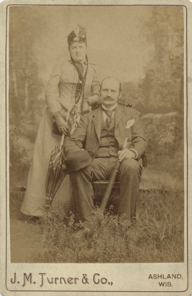 Studio portrait in front of a painted backdrop of a woman and man posing together. The man is sitting in a chair holding a hat and an umbrella. The woman is standing next to him leaning on the back of the chair, and holding an umbrella. There is grass in the foreground, and the painted scene behind them is of foliage and a stone wall, so it appears they are posing outdoors.