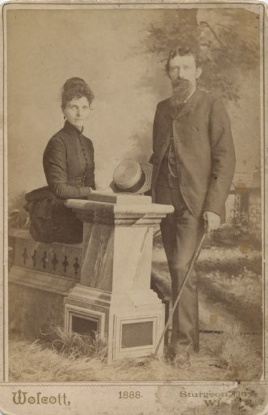 Studio portrait in front of a painted backdrop of a woman and man posing together. The woman is wearing a dark-colored dress and is sitting on a prop stone balustrade and leaning her arm on the column. The man is standing on the right near the column, and is holding a straw hat and a cane.