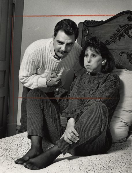 View of a woman sitting on a bed with her knees bent, propped up by pillows. A man is standing next to her holding her hand. The woman appears to be holding her breath. Caption reads: "Ed and Linda Krueger practice childbirth exercises."