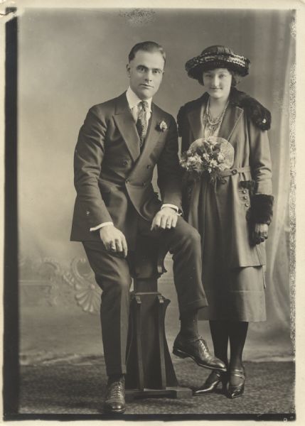 Studio portrait in front of a painted backdrop of a man and a woman posing together. The man is half-sitting on a pedestal, and is wearing a suit with a boutonniere in his lapel. The woman is standing and is wearing a hat, overcoat, dress and gloves, and is holding a bouquet of flowers.
