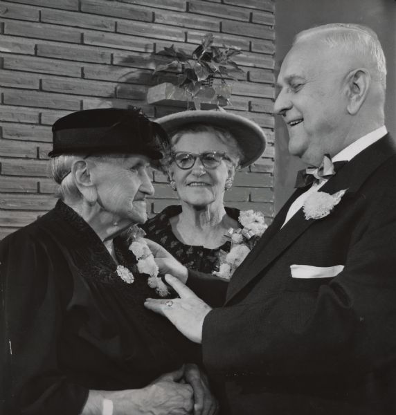 A man is pinning a corsage onto the lapel of an older woman, while another woman is looking on. Caption reads: "<b>The 93 year old mother</b> of one of the principals was present Monday when Mr. and Mrs. Joseph Schultz, both 73, of 1645 S. 29th St., celebrated their 50th wedding anniversary. Schultz pinned a corsage on his mother, Mrs. Wilhelmina Schultz, 3055 S. 17th St. The party was held at Tuckaway Country Club."