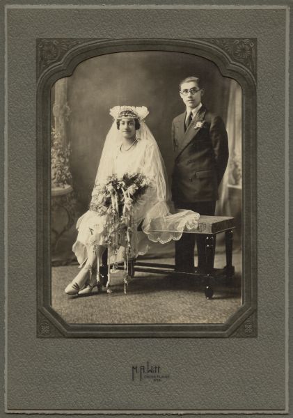 Studio portrait in front of a painted backdrop of a woman and man posing together. The woman is wearing a bridal gown with veil, and is sitting on a bench and holding a bouquet of flowers. The man is standing beside her on the right, and is wearing a suit and boutonniere in his lapel. Caption reads: "Mr. and Mrs. Leo Meicher."