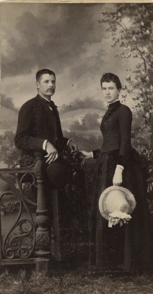 Studio portrait in front of a painted backdrop of a man and woman posing together on either side of a prop wrought-iron fence. The man is wearing a suit and holding a hat. The woman is wearing a dark-colored dress and is holding a straw hat in one hand.