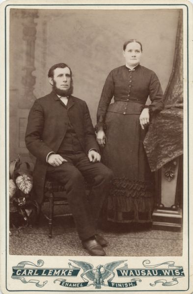 Studio portrait in front of a painted backdrop of a man and woman posing together. The man is wearing an overcoat and suit and is sitting in a chair. The woman is wearing a dark-colored dress and is standing and leaning on a pedestal that is covered by drapery.