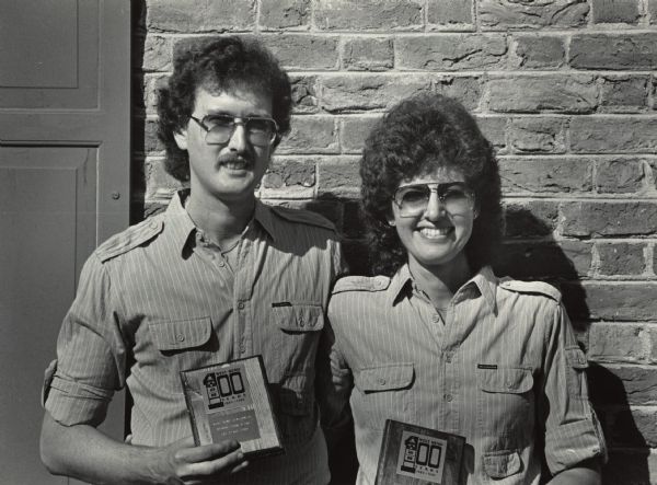 Waist-up portrait of a man and woman posing together outdoors while standing against a brick wall. They are both are wearing matching sunglasses and shirts, and they have similar hairstyles. They are holding plaques that read: "West Bend 100 Years 1885-1985, West Bend Centennial Spouse Look-Alike First Place 1985." Caption reads: "Jeff and Pat Otten, winners of the spouse look-alike contest."