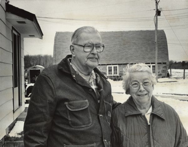 Outdoor portrait of a man and woman posing together. They are both wearing jackets, and snow is on the ground, and a barn is in the background. Caption reads: "Frank and Louise Rombalski remember what it was like before electricity came to rural America."