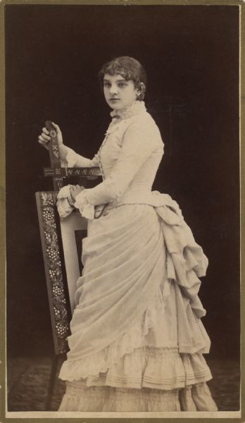 Portrait of a woman posing in a studio, leaning on what appears to be a framed picture on a decorated easel.