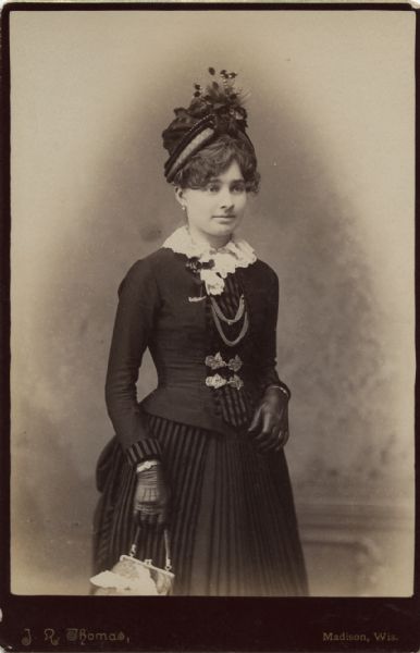 Three-quarter length portrait of a woman standing in front of a backdrop. She is holding a handbag and is wearing gloves, a dark-colored dress, and a hat.