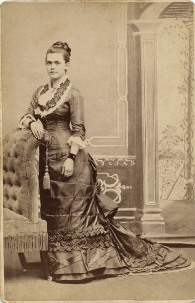 Full-length portrait in front of a painted backdrop of a woman posing in  while leaning on an upholstered piece of furniture with a tassel. The woman is wearing a dress with elaborate ruffles and a train, a necklace, and dark cuffs on each wrist.