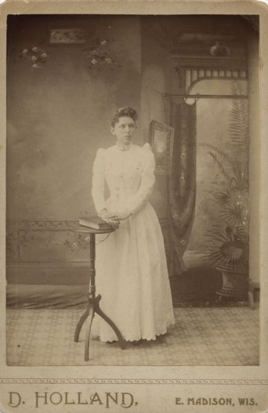 Full-length studio portrait in front of a painted backdrop of a woman standing and resting her hands on a book, which is on a small table. She is wearing a white floor length dress with puffed sleeves.
