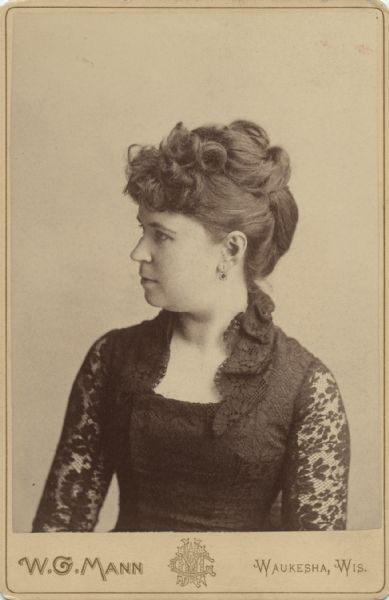 Waist-up studio portrait of a woman with her head turned to the left in profile. She is wearing a dark-colored lace dress.