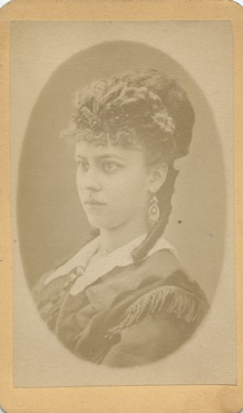 Oval-framed quarter-length portrait of a woman. She is wearing earrings, a dress with fringe at the shoulders, and a tiara. She has a pompadour hairstyle with long curls.