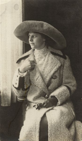 Portrait of a woman sitting next to lace-curtained window. She is wearing a hat with a large brim and a long coat. She has a glove on one hand, which is on her lap holding the other glove.