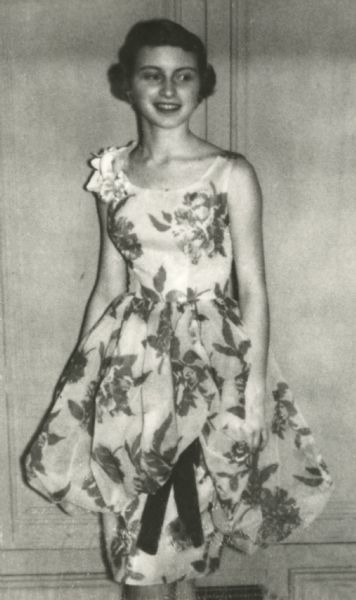 Laura Foster (b. 1941) standing in the foyer of her house dressed for her senior prom at Rockford East High School. The sleeveless, knee-length dress is white with a floral design and has a bubble or balloon overskirt and a narrow underskirt. There is a dark ribbon bow attached to the underskirt at the proper left. There is a floral corsage at her proper right shoulder.