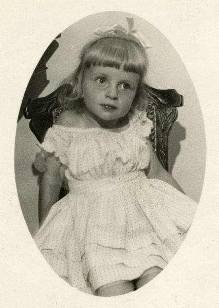 Portrait of 2-year old Laura Foster (b. 1941) sitting in a dark-colored, highly-carved chair. She is wearing her maternal great-grandmother's girlhood dress from 1862-1863. The dress is white with small polka dots. It is knee-length, off-the-shoulder, and has two horizontal tucks near the skirt hem and a puffy bodice. Anne also has a hair bow in her blonde hair.

Laura's great-grandmother was Mary Adell "Della" (Waldo) Henderson (1858-1939). Della wore this dress in Winneconne (Winnebago County), Wisconsin.

The photograph was taken in Rockford, Illinois or Green Bay, Wisconsin.