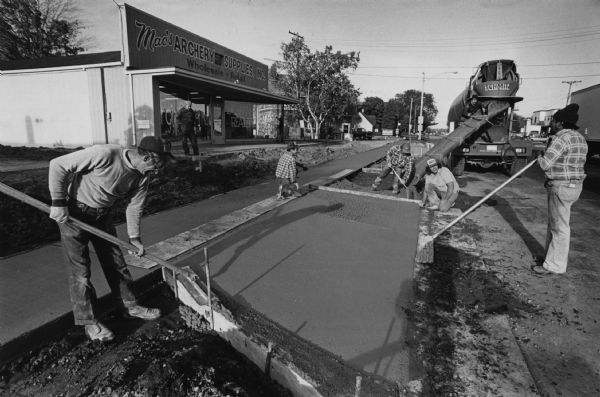 A group of five people are working on freshly poured cement outside a shop called Mac's Archery Supplies, Inc. Caption reads: "A group from Wiggins Construction Co., Menomonee Falls, worked on a driveway entrance in Saukville."