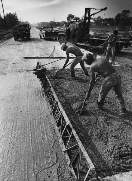 Two shirtless men are standing in freshly poured cement using shovels. A third man is standing behind them. Caption reads: "Jim Kreilkamp (left) and Roy Heldt, employes of Cape Construction Co., spread concrete into forms on Mequon Rd., where crews were pouring new intersections. Finished sections were behind the workers, who were at Oriole La. and Mequon Rd. in Mequon."