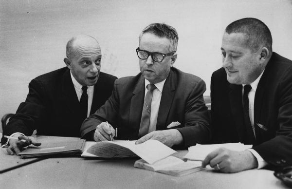 Three men are sitting together at a table. The man in the middle appears to be writing on a document, while the man on his right is talking to him. Caption reads: "James T. Horaitis, business agent of local 317 of the operating engineer's union (center), carefully checked documents pertaining to the tentative agreement to end the 11 day old Electric Co. strike. At the left was Robert E. Graz, the union attorney. Thomas J. Cassidy (right), was the Electric Co. negotiator."