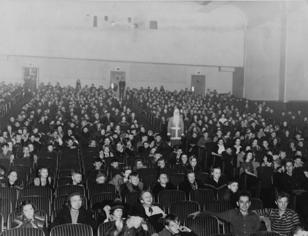 View from the front of a theater towards children sitting in the audience. Standing in the aisle on the right is a man who is dressed as Santa Claus.