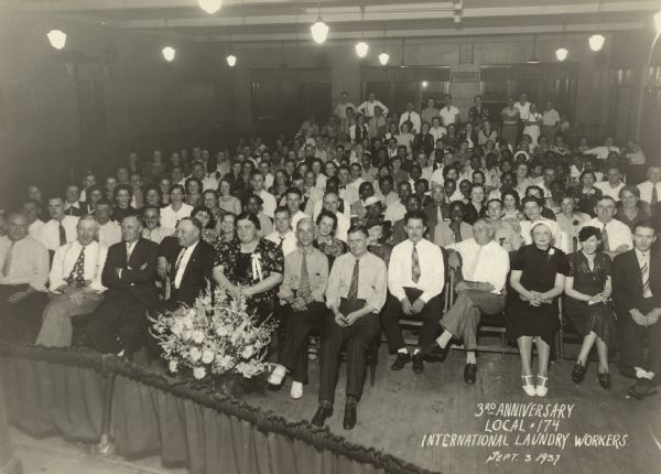 View of a large group of people sitting and standing in a large room. A floral bouquet and a curtain divider are in the foreground.