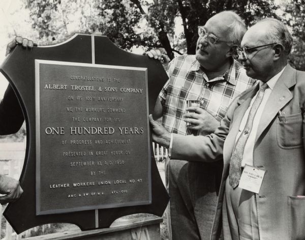 Two men are standing outdoors and holding onto a plaque, which reads: "CONGRATULATIONS TO THE ALBERT TROSTEL & SONS COMPANY ON ITS 100TH ANNIVERSARY. WE, THE WORKERS, COMMEND THE COMPANY FOR ITS ONE HUNDRED YEARS OF PROGRESS AND ACHIEVEMENT. PRESENTED IN GREAT HONOR ON SEPTEMBER 13, A.D. 1958 BY THE LEATHER WORKERS UNION LOCAL NO. 47. AMC & BW OF N.A. AFL-CIO."<p>Image caption reads: "<b>A plaque</b> honoring a Milwaukee firm's 100th anniversary was presented by a labor union. The award was made at a picnic for employees of the Albert O. Trostel & Sons Co. at the Old Heidelberg Park, 5423 N. Port Washington Rd., Glendale. From left are Albert Trostel, Jr., company president and John Churka, secretary-treasurer of local 47 of the Amalgamated Meat Cutters and Butcher Workmen (AFL-CIO). The plaque was mounted on a board in the shape of a hide."</p>