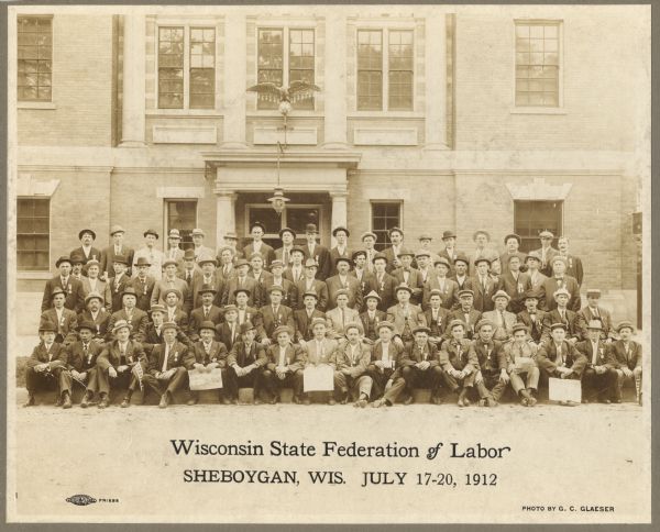 Group portrait of a large group of men who are wearing suits and hats posing in front of a building. The men are also wearing medals on their lapels.