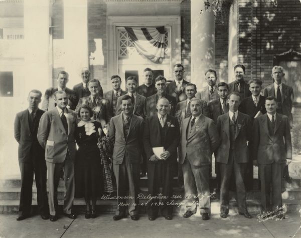 Group portrait of 22 people posing in front of a building. A caption on the back of the image identifies many of the people, though it does not seem to account for everyone. It reads: "Wisconsin Delegation, 56th A.F. of L. Convention. Nov. 16-27, 1936. Tampa, Florida. Front row left to right: Felix Olkines, Kenosha; Mabel Melvin, Wausau; Emil Costello, Kenosha; Stanley Joers, Milwaukee; Joseph A. Padway; Erwin Zumach, Milwaukee; Steve J. Thomas, Racine. Second row left to right: August J. Scherr, Milwaukee; Cedric Parker, Madison; Andrew B. Cross, Kenosha; Joseph M. Driscoll, Milwaukee; David Sigman, Two Rivers; Robert W. Powers, Port Washington; Rudolph Faupl, Milwaukee. Third row left to right: Charles Heymanns, Sheboygan; John J. Handley, Miklwaukee; J. F. Friedrich, Milwaukee; Father Francis J. Haas, Milwaukee; Arnold S. Zander, Madison; Henry Rutz, Milwaukee; Paul Porter, Kenosha; and William H. Sommers, Racine."