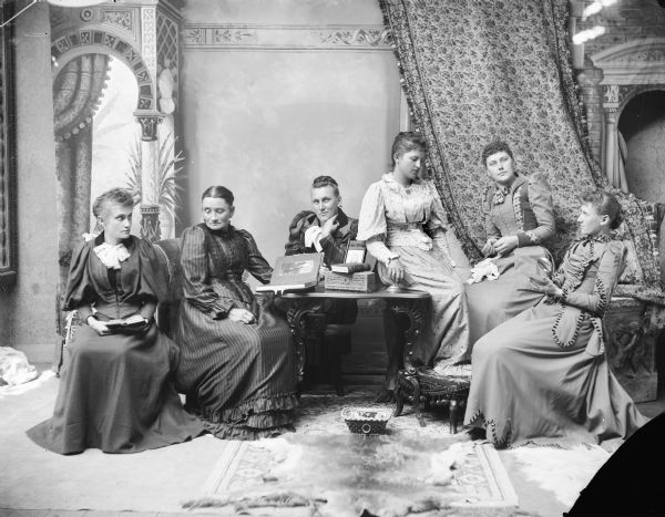 Studio portrait in front of a painted backdrop, and a draped tapestry or cloth, of six women, sitting or standing around a table. One woman at the table appears to be holding a photograph album. A woman sitting on the right is sewing, and the woman on the far right is holding a magnifying glass in her right hand to look at something she is holding in her left hand. There is a fur rug on the floor.